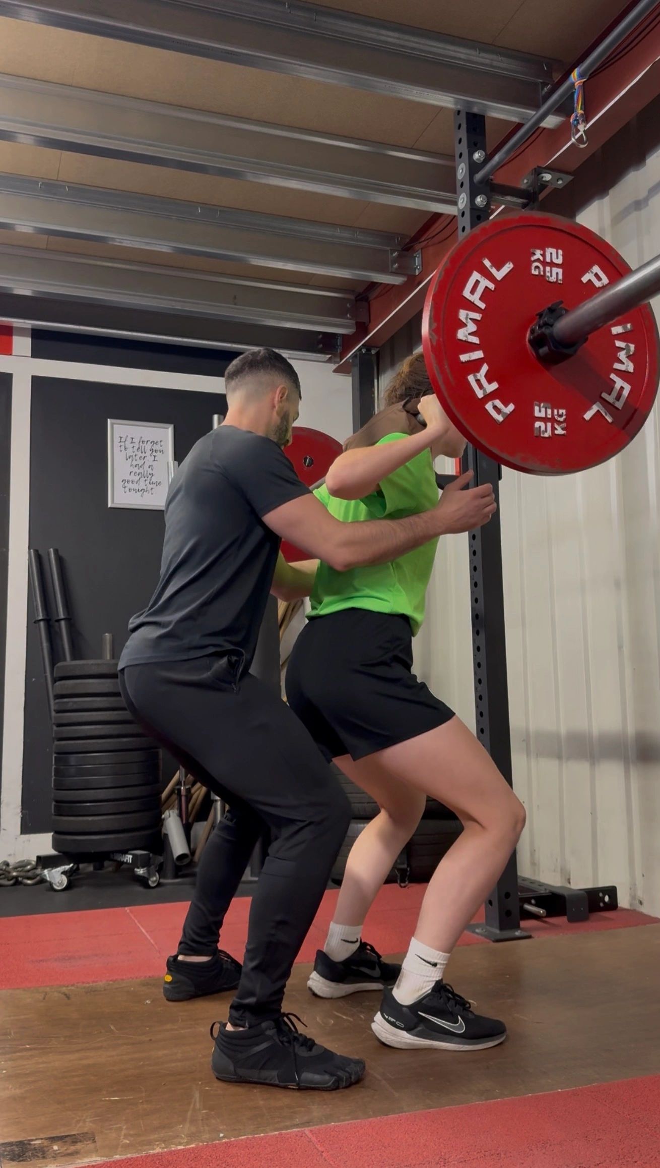 Alexandros in black training clothes supporting a woman during a barbell squat